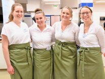4 Personen mit weißen Oberteilen und grünen Schürzen stehen in einem Raum nebeneinander